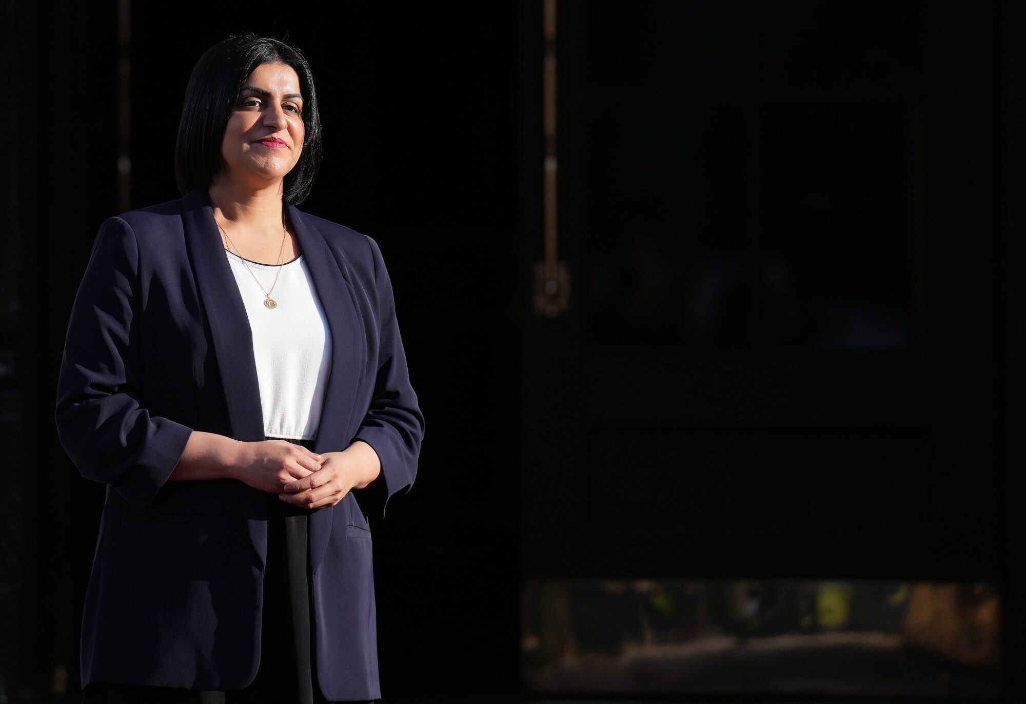 Shabana Mahmood stood to the left, facing the camera, with a dark background behind her