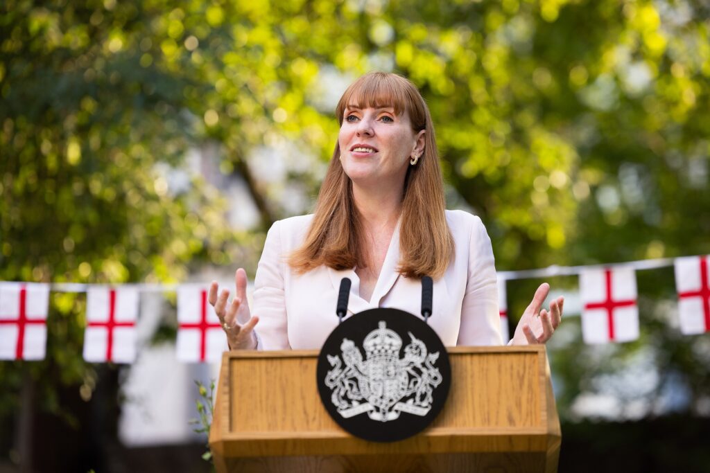  Angela Rayner standing behind a podium, with English flags in the background, looking up and off the to side