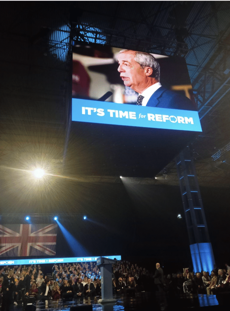Nigel Farage, Reform UK leader, on a large projecter board in the air, with the podium below it.